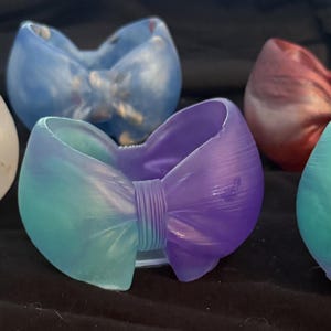 May include: Six resin bow-shaped containers in various colors. The colors include white with gold flecks, blue with gold, red with gold, teal and purple, and teal and blue. The containers are on a black surface.