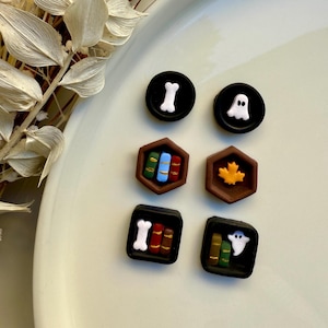 Puede incluir: Una colección de botones decorativos con varios diseños. Los botones incluyen un hueso blanco, un fantasma, una hoja de arce y estanterías en miniatura con libros coloridos. Los botones son negros, marrones y blancos.