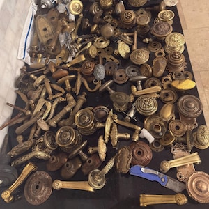 May include: A collection of antique brass door handles, knobs, and hardware. The assortment includes various shapes and sizes, with ornate detailing and aged patinas. The hardware is arranged on a dark surface, showcasing the variety of vintage designs.