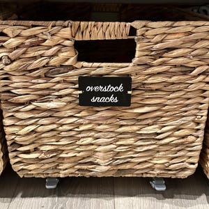 May include: Woven wicker storage basket with a rectangular shape and a cut-out handle. A small black label reads "overstock snacks". The basket is light brown and sits on small gray wheels.