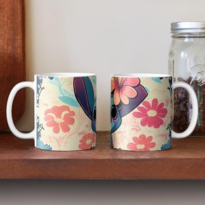 May include: Two white ceramic mugs with a floral pattern and a cartoon character with blue fur and a flower crown. The character is on a light yellow background with pink and blue flowers.