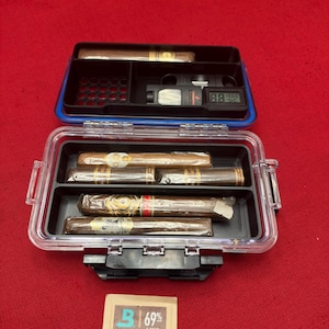 May include: A clear plastic travel humidor case containing five cigars, a black cigar cutter, and a digital hygrometer. Below the case lies a tan Boveda 69% 2-way humidity control packet with the text 69% and No Mess. No Worries.