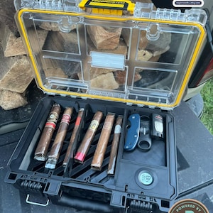 May include: A black and clear plastic storage case with a yellow latch, containing six cigars in individual wrappers, a blue folding knife, a black cigar cutter, a black lighter, and a round digital hygrometer with a Boveda humidity control logo.