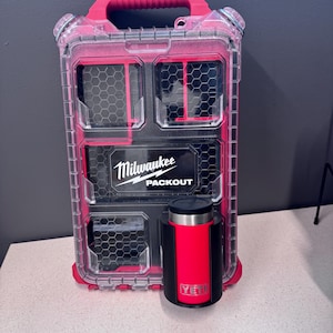 May include: A clear and pink Milwaukee Packout storage container with a black and red Yeti tumbler. The container has multiple compartments with a honeycomb pattern. The Yeti tumbler is red and black.