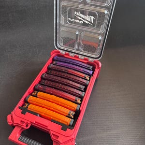 Peut inclure: Une mallette de rangement rouge Milwaukee Packout avec un couvercle transparent, ouverte pour révéler plusieurs accessoires d'outils orange, violets et rouges. La mallette a une poignée et un loquet. Le logo Milwaukee est visible sur le couvercle.