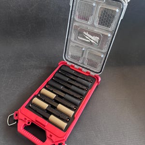 May include: Open red and clear plastic storage case with multiple compartments. The case contains several black and tan cylindrical objects. The clear lid has the brand name "Milwaukee" printed on it.