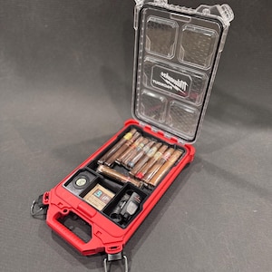 May include: A red, open, rectangular case with a clear lid, containing cigars, a small black container, and other accessories. The case has a handle and metal loops. The cigars are various shades of brown. The case is on a dark surface.