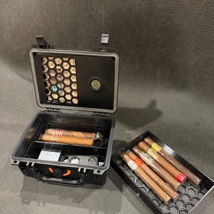 May include: A black, rectangular, hard-shell travel humidor case with a latch, featuring an integrated digital hygrometer and a honeycomb-patterned interior tray. The case contains two cigars, while a separate matching tray holds four additional cigars.