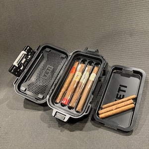 May include: A black YETI hard-shell case with a hinged lid, featuring a textured interior and a molded tray holding four cigars. A separate black YETI lid rests nearby, with three additional cigars placed horizontally across its surface.