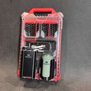 May include: A red and clear plastic tool organizer with a red handle. It contains a black Stanley flask and a green Stanley thermos. The organizer has the "Milwaukee PACKOUT" logo.