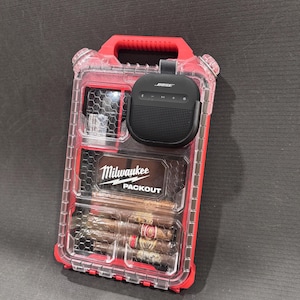 May include: A red and clear Milwaukee Packout storage case with a black Bose speaker attached. The case contains cigars and other items. The Milwaukee Packout logo is visible on the front.