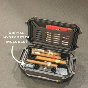 May include: A black, rectangular, hard-shell travel case with an open lid, containing four cigars, a silver metal lighter, and a built-in digital hygrometer displaying 63%. The lid features a black grid panel with text reading DIGITAL HYGROMETER INCLUDED!