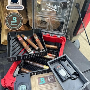 May include: A red and black Milwaukee Packout storage case containing multiple cigars, a Boveda 69% humidity control pack, a digital hygrometer, and a black cigar cutter. The clear lid features a hexagonal pattern and the Milwaukee logo.