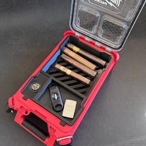 May include: Red Milwaukee Packout humidor with a clear lid, holding cigars, a cigar cutter, and a lighter. The humidor has a digital hygrometer displaying 42%. The lid is open, revealing the contents.