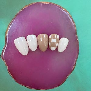 May include: Five fake nails on a pink and gold agate coaster. Two nails are white, one is brown, and two are white with a brown checkered pattern.
