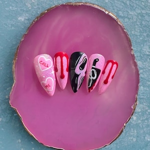 May include: Pink acrylic nails with various designs. One nail has the text "CALL ME" on a heart. Other nails feature dripping blood, a black and white ghost face, and a knife. The nails are displayed on a pink agate slice.