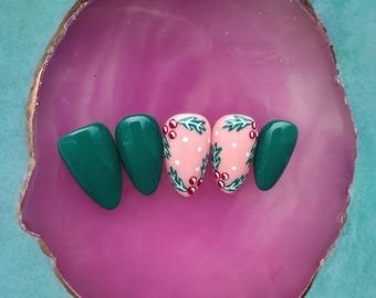 Puede incluir: Cinco uñas postizas, dos son verdes y tres son rosas con un diseño de acebo blanco y bayas rojas.