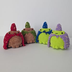 May include: Four handmade crochet frog plushies in various colors. Each frog wears a cape and a pointed hat, with details like yellow cross stitches. The frogs are brown, green, blue, and lime green.
