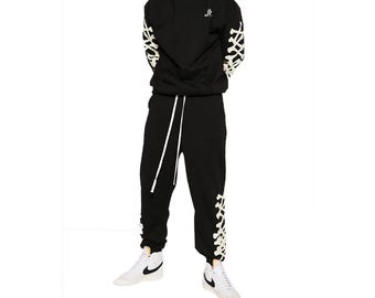 May include: Black hoodie and sweatpants set with white accents. The hoodie features a small logo on the chest and decorative white patterns on the sleeves. The sweatpants have a drawstring waist and matching white patterns on the lower legs. The set is paired with white sneakers.