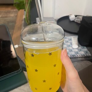 Puede incluir: Un vaso de vidrio transparente con tapa y pajita, lleno de una bebida amarilla. El vaso tiene un patrón de estrellas doradas en el exterior.