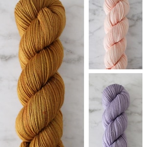May include: Three skeins of yarn in different colors: a deep golden yellow, a pale pink, and a light lavender.