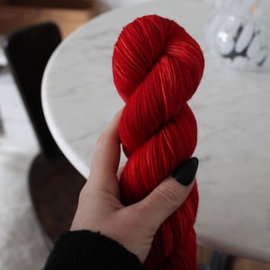 May include: A skein of red yarn with white highlights. The yarn is wound into a ball and held in a hand.