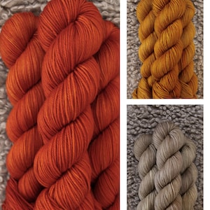 以下が含まれることがあります： オレンジ、ゴールド、グレーの3色の糸の玉。糸は柔らかくふわふわしていて、編み物や編み物に最適です。