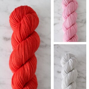 May include: Three skeins of yarn in different colors: red, pink, and gray. The yarn is wound into a ball shape and is ready to be used for knitting or crocheting.