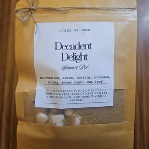 May include: A brown paper pouch tied with twine at the top, containing a simmer pot mix. The white label reads: STACE AT HOME, Decadent Delight, Simmer Pot, marshmallow, cocoa, vanilla, cinnamon, nutmeg, brown sugar, bay leaf, and instructions.