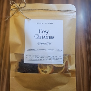 Könnte beinhalten: Ein brauner Papierbeutel, der mit einer Schnur verschlossen ist und eine Cozy Christmas Simmer Pot Mischung enthält. Das Etikett listet Cranberry, Zimt, Orange und Muskatnuss auf, mit Anweisungen, den Inhalt in 710 ml kochendes Wasser zu geben.