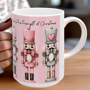 May include: White ceramic mug with a pink background and three illustrated nutcracker figures. The text "Love is the true gift of Christmas" is printed on the mug.