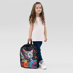 May include: A black backpack featuring a colorful cat design with a splash of vibrant paint. The cat has multicolored eyes and fur. The backpack has a starry pattern and a top handle.