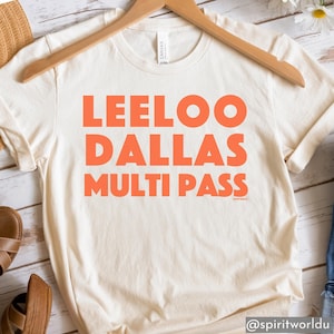 May include: Cream-coloured t-shirt with the words "LEELOO DALLAS MULTI PASS" in large orange letters. The shirt is hanging on a wooden hanger.