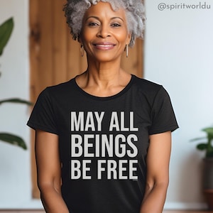 Puede incluir: Camiseta negra de cuello redondo con el texto blanco "MAY ALL BEINGS BE FREE". La camiseta está hecha de un material suave. La imagen muestra a una persona que lleva la camiseta.
