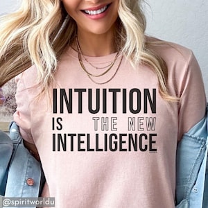 May include: A pale pink t-shirt featuring the statement "INTUITION IS THE NEW INTELLIGENCE" in bold black text. The shirt is paired with a denim jacket and a gold necklace. The image is well-lit.
