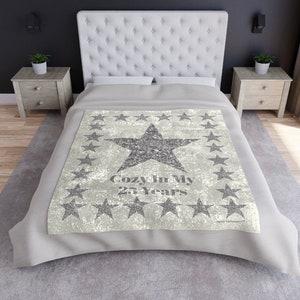May include: A light gray blanket with a large, glittery star in the center, surrounded by smaller stars. The text "Cozy In My 25 Years" is printed below the star. The blanket is on a bed with a white headboard.