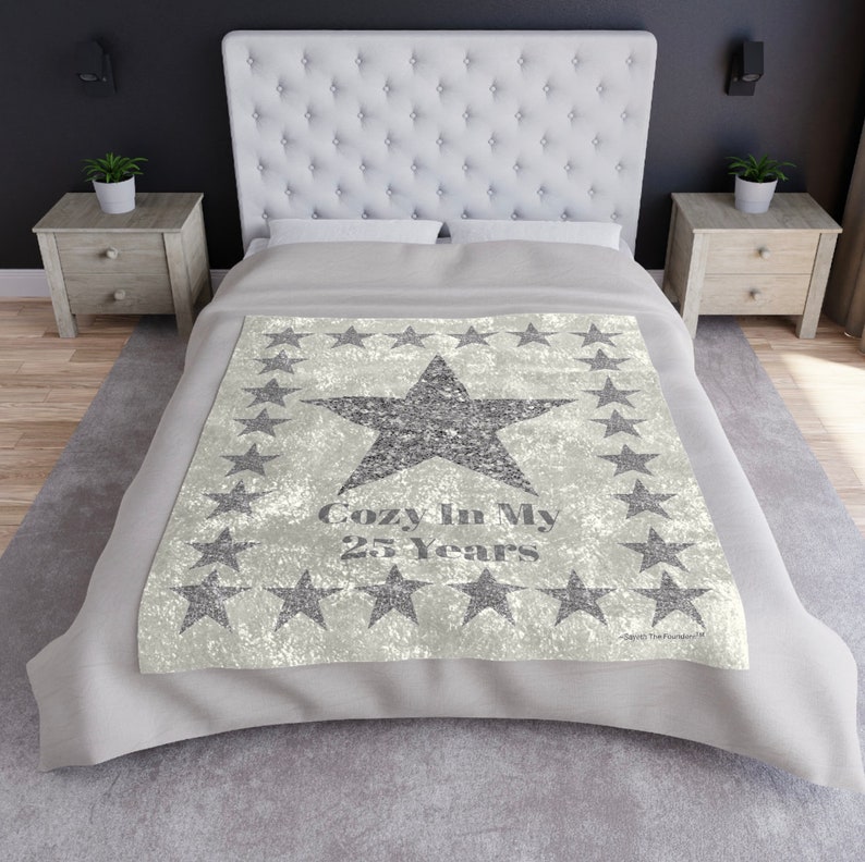 May include: A light gray blanket with a large, glittery star in the center, surrounded by smaller stars. The text "Cozy In My 25 Years" is printed below the star. The blanket is on a bed with a white headboard.