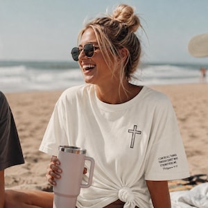 Puede incluir: Una camiseta de manga corta en color blanco roto que presenta un gráfico de una cruz negra en el pecho izquierdo y el texto "I CAN DO ALL THINGS THROUGH CHRIST WHO STRENGTHENS ME. Philippians 4:13" impreso en negro en la manga izquierda.