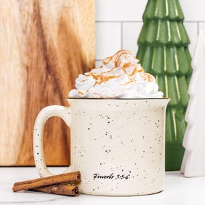 May include: A cream-coloured ceramic mug with black speckles, featuring the text "Proverbs 3:5-6" in black script. The mug is filled with whipped cream topped with cinnamon and is positioned next to two whole cinnamon sticks on a white surface.