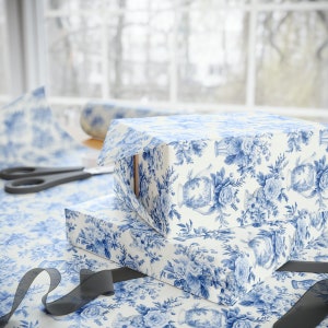 Könnte beinhalten: Zwei Geschenkboxen, die in weißes und blaues Blumenmusterpapier eingewickelt sind. Die Boxen sind übereinander gestapelt. Die obere Box ist leicht geöffnet und zeigt ein braunes Inneres.