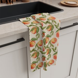 May include: A kitchen towel with a repeating pattern of watercolor peaches and green leaves on a cream-colored background. The towel is draped over the edge of a countertop and sink, showcasing the fruit-themed design. The towel is made of a textured fabric.