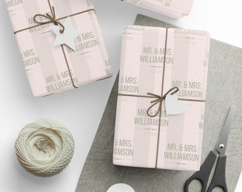 Pode incluir: Papel de embrulho listrado rosa e branco com o texto "MR. & MRS. WILLIAMSON EST. 2024" repetido no papel. O papel está embrulhado em torno de uma caixa de presente com um laço de barbante marrom e uma etiqueta branca em forma de coração.