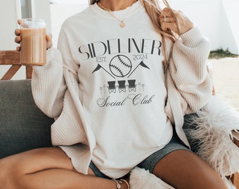 May include: Cream-colored sweatshirt with the text "SIDELINER EST. 2024 Social Club" and a baseball graphic. The sweatshirt is worn with denim shorts and a cream-colored cardigan. A glass of coffee is held in the hand.