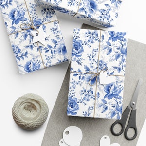 Op de afbeelding: Drie cadeaudozen verpakt in blauw en wit bloemmotiefpapier met touw en papieren etiketten. De dozen staan op een grijs linnen oppervlak met een bol touw en een schaar.