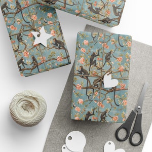 Puede incluir: Regalos envueltos con papel de regalo con estampado de monos y flores. El papel turquesa presenta monos, flores rosas y ramas. Los regalos están atados con hilo y adornados con etiquetas blancas en forma de estrella y corazón. También se ven un ovillo de hilo y unas tijeras.