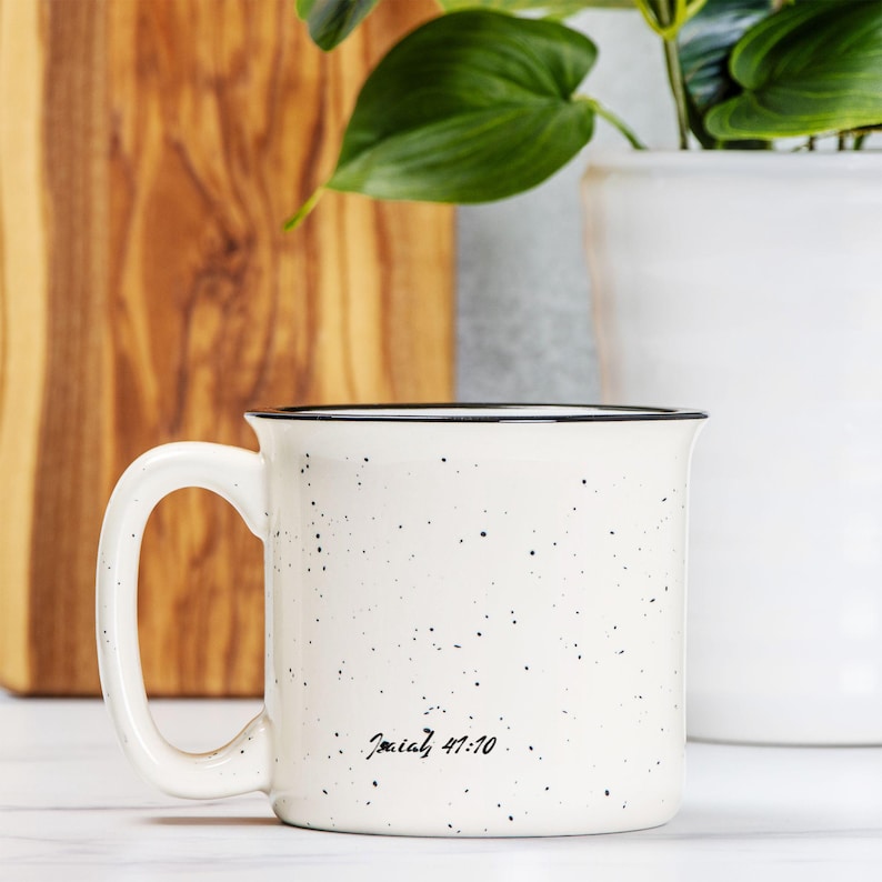May include: A cream-coloured ceramic mug with a speckled finish and a black rim. The mug features a handle on the left side and the black printed text Isaiah 41:10 near the bottom. The design is simple with a smooth, glossy surface texture.