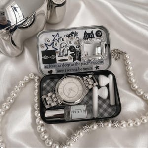 May include: A silver wristwatch with a white face and a matching metal band sits inside a small, open, black and white checkered tin. The tin also contains white earbuds, a small perfume bottle, and a lipstick. The tin is decorated with stickers and the text "at least as deep as the pacific ocean now i wanna be yours".