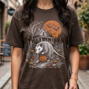 May include: A dark brown short-sleeve t-shirt featuring a graphic illustration of an opossum holding a jack-o'-lantern bucket. The scene includes a house, a large orange moon, bats, and autumn leaves, rendered in a detailed, sketch-like style.