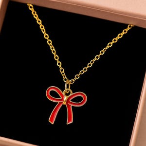 May include: A gold-colored necklace with a red bow pendant, presented in a pink gift box. The bow pendant has a red enamel finish with gold-colored outlines. The necklace chain is also gold-colored.