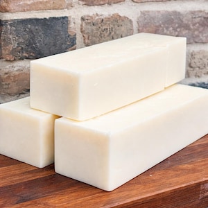 May include: Three rectangular bars of off-white soap are stacked on a wooden surface. The soap bars have a smooth, solid appearance. The background features a brick wall, adding a rustic touch to the composition.